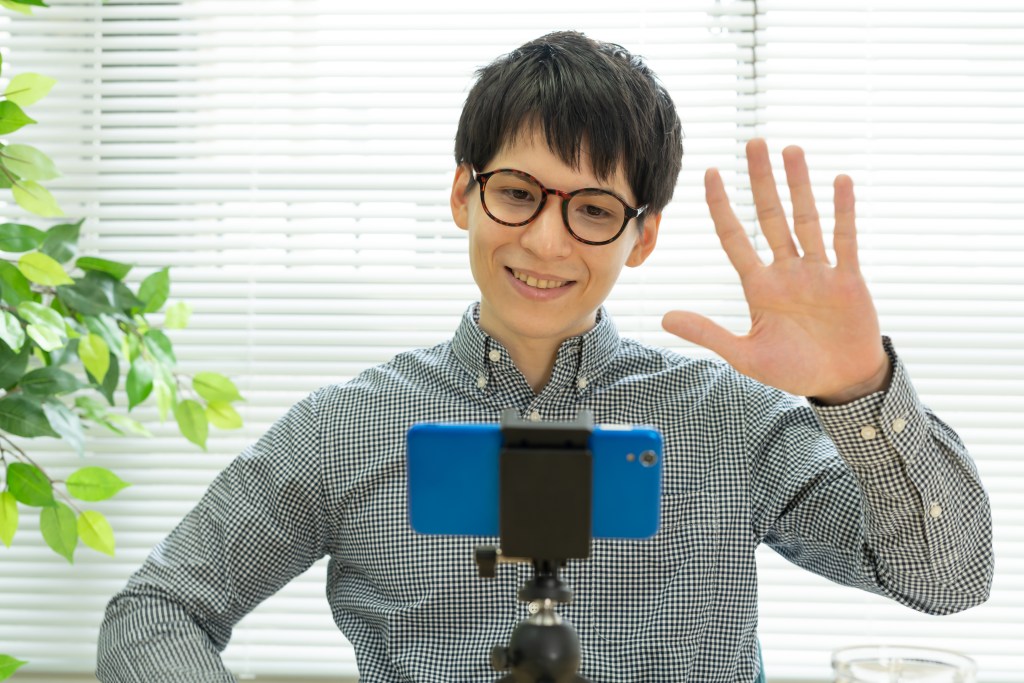 カメラに向かって手を振る男性。チェック柄のシャツを着て、眼鏡をかけている。背景には植物とブラインドがある。