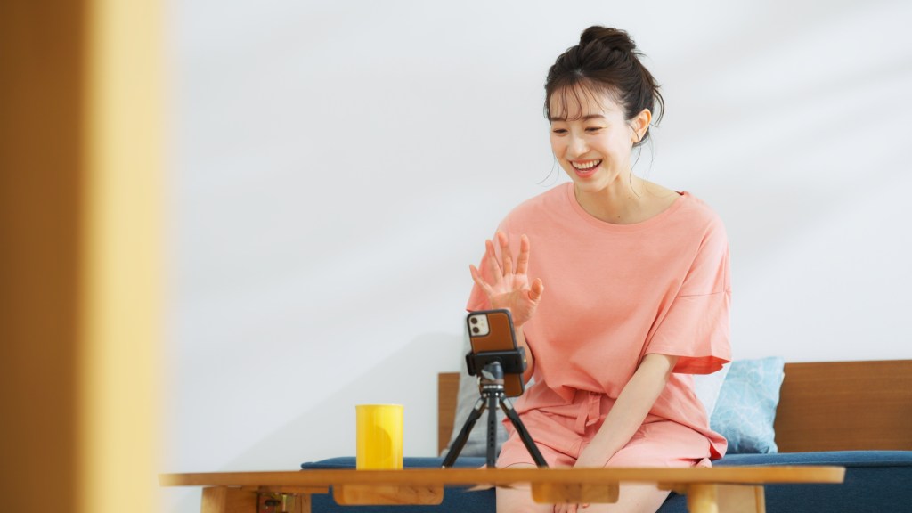 カメラに向かって手を振る女性。彼女はピンクのトップスを着ており、テーブルの上にはスマートフォンと黄色いカップが置かれている。